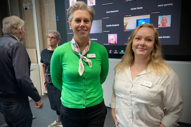 Två kvinnor framför två andra mötesdeltagare och en skärm med onlinedeltagare.