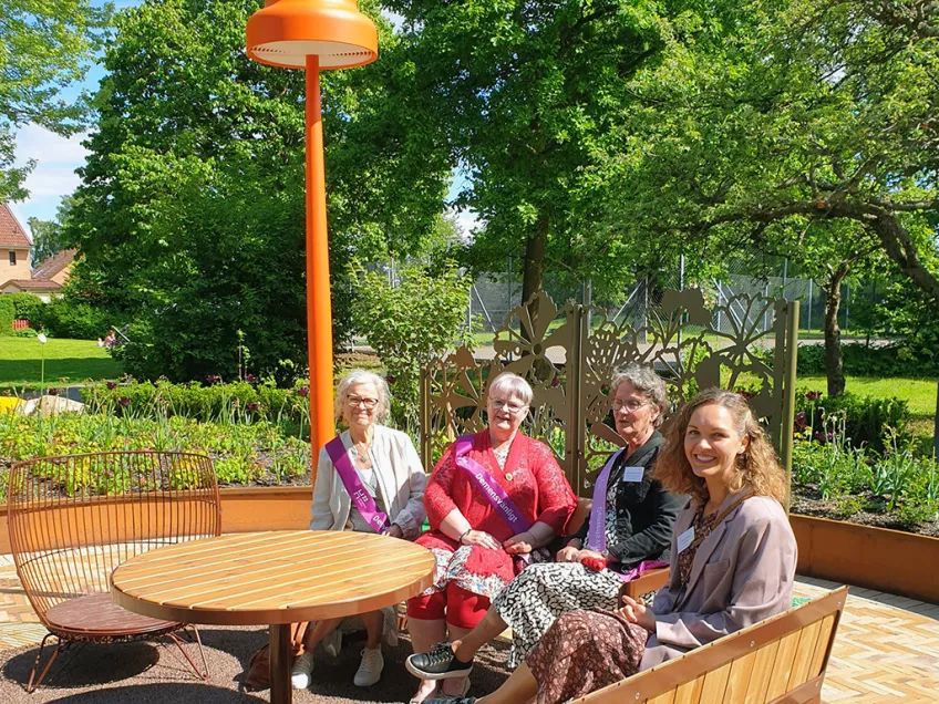 Fyra kvinnor sitter på utemöbler i trä, sidan av stor orange golvlampa, i solig, grön park.